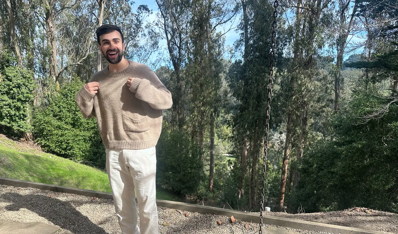 A man wearing a beige knitted crewneck sweater from soft in an outdoor setting.